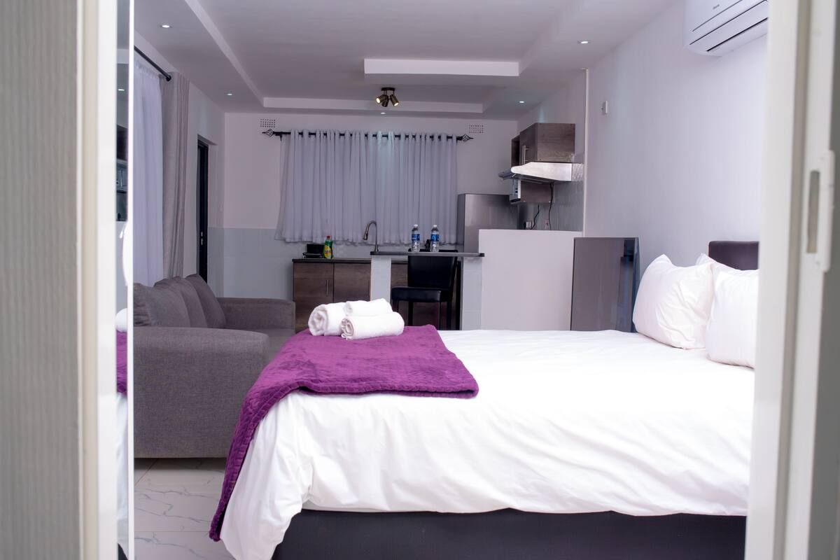 A modern hotel room with a bed, a sofa, a desk with a chair, and a kitchenette.