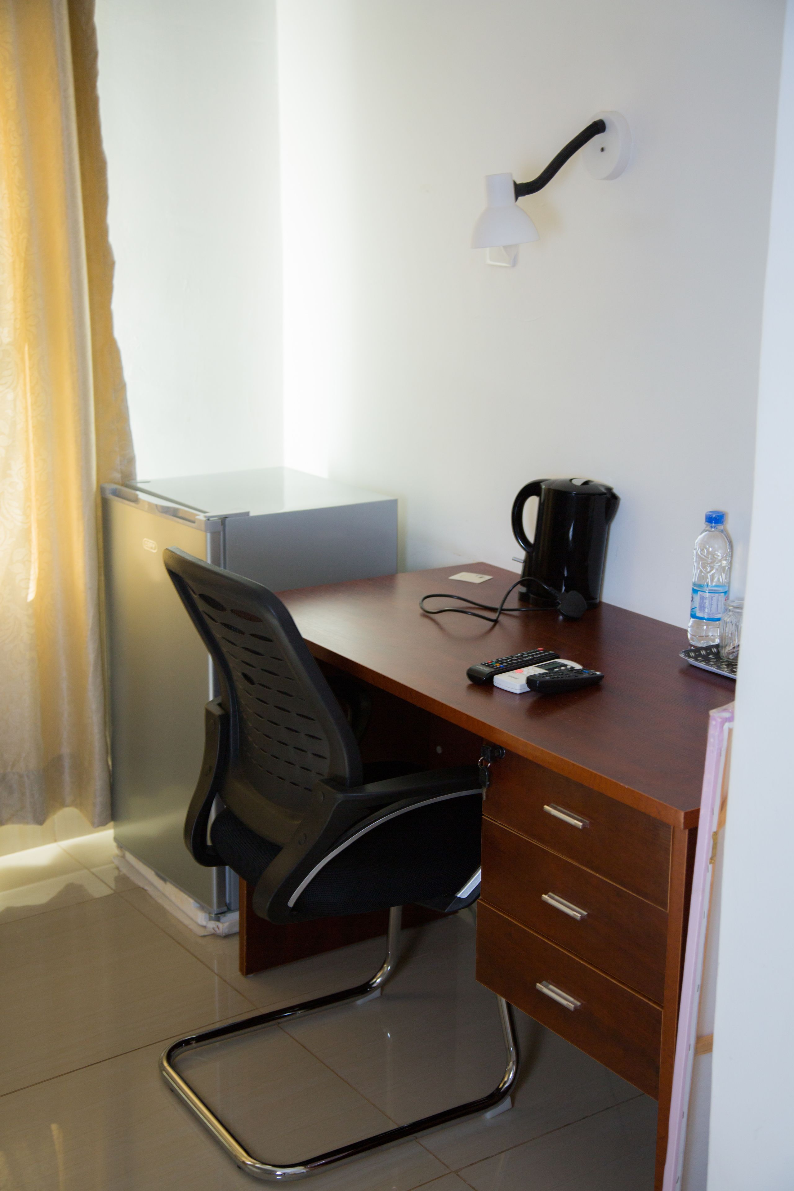 A compact hotel room with a wooden desk, a black office chair, a coffee maker, a bottle of water, and a remote control.