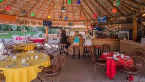 An outdoor restaurant with thatched roof, tables with yellow and red tablecloths, and a bar area.