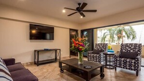 A living room with a sofa, a coffee table with a vase of flowers, patterned armchairs, a ceiling fan, and a TV mounted on the wall.