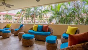 A balcony with wicker furniture and blue cushions, overlooking palm trees and a building with a red roof.