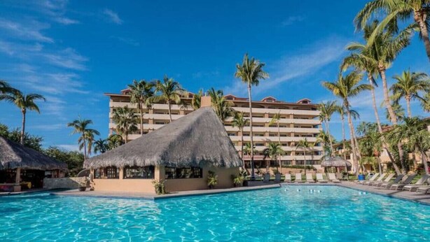 A resort pool with a thatched-roof structure, palm trees, and a multi-story building in the background.