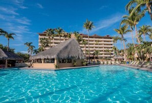 A resort pool with a thatched-roof structure, palm trees, and a multi-story building in the background.