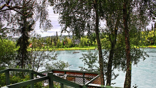 Eine Holzterrasse mit Blick auf einen Fluss, im Hintergrund Bäume und Häuser.