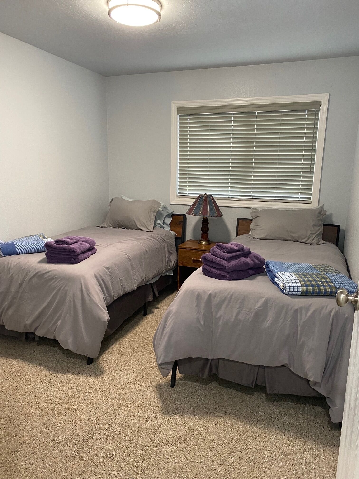 A bedroom with two beds, a nightstand, a lamp, and a window with blinds.