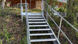 Eine Holzhütte mit Steinfundament, einer Metalltreppe, die zu einer Holzterrasse führt, und umliegenden Bäumen.