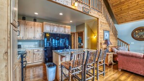 A spacious kitchen with wooden cabinets, a central island with bar stools, a refrigerator, and a dining area with a sofa and lamp.