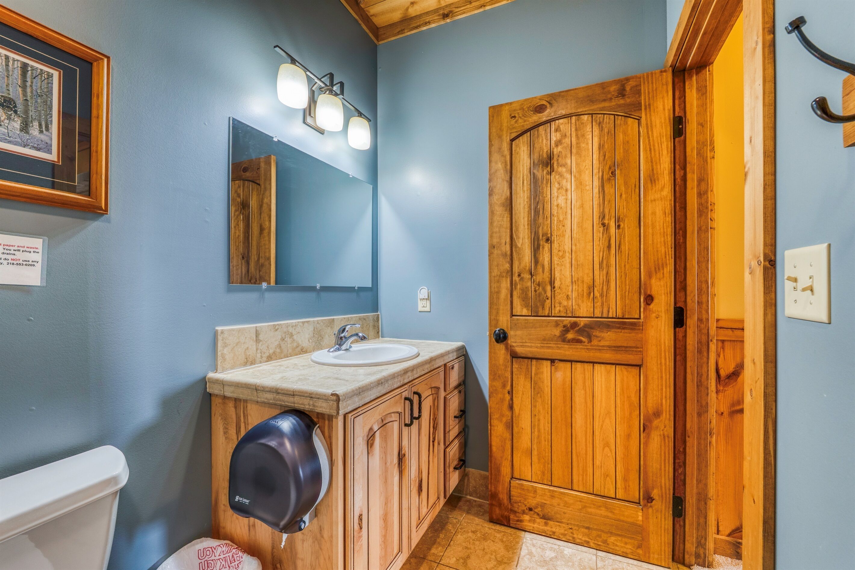 A bathroom with a wooden door, a sink, a mirror, and a towel dispenser.