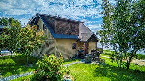 A log cabin with a porch and a small deck, surrounded by a grassy yard and trees.