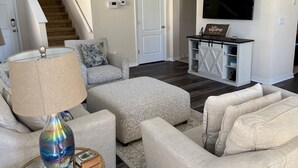 Un salon avec un ensemble de canapés, une table basse avec une lampe, un téléviseur fixé au mur et un escalier en arrière-plan.