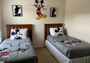 Two twin beds with Mickey Mouse-themed bedding, a wall with Mickey Mouse artwork, and a plain wall.
