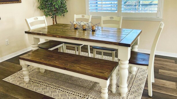 Un espace repas comprenant une table en bois, des chaises blanches, un banc et un tapis à motifs.