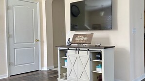 Un espace d’accueil avec une porte blanche, un téléviseur à écran plat fixé au mur et un panneau d’accueil placé au-dessus d’une console blanche dotée d’étagères.