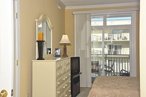 A hotel room with a bed, a dresser, a mirror, a lamp, and a view of a balcony with railings.