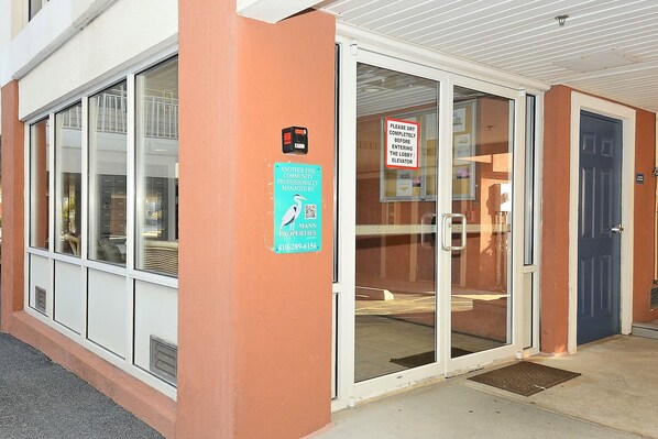 A building entrance with a glass door, a sign indicating a fine community professionally managed by Mann Properties, and a security device on the wall.