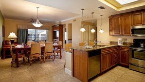A spacious kitchen with wooden cabinets, a stainless steel oven, and a granite countertop. There is a dining area with wicker chairs and a table, and a large window providing natural light.