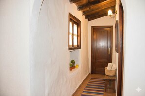 Un pasillo con una puerta de madera, un pequeño estante con plantas, un banco y una alfombra a rayas de colores.