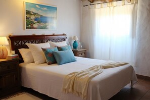 A neatly made bed with a white bedspread and multiple pillows, a wooden headboard, a bedside table with a lamp, and a painting of a coastal scene on the wall.