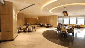 A spacious hotel dining area with multiple tables and chairs, a curved black marble floor, and a modern ceiling design with recessed lighting.