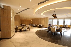 A spacious hotel dining area with multiple tables and chairs, a curved black marble floor, and a modern ceiling design with recessed lighting.