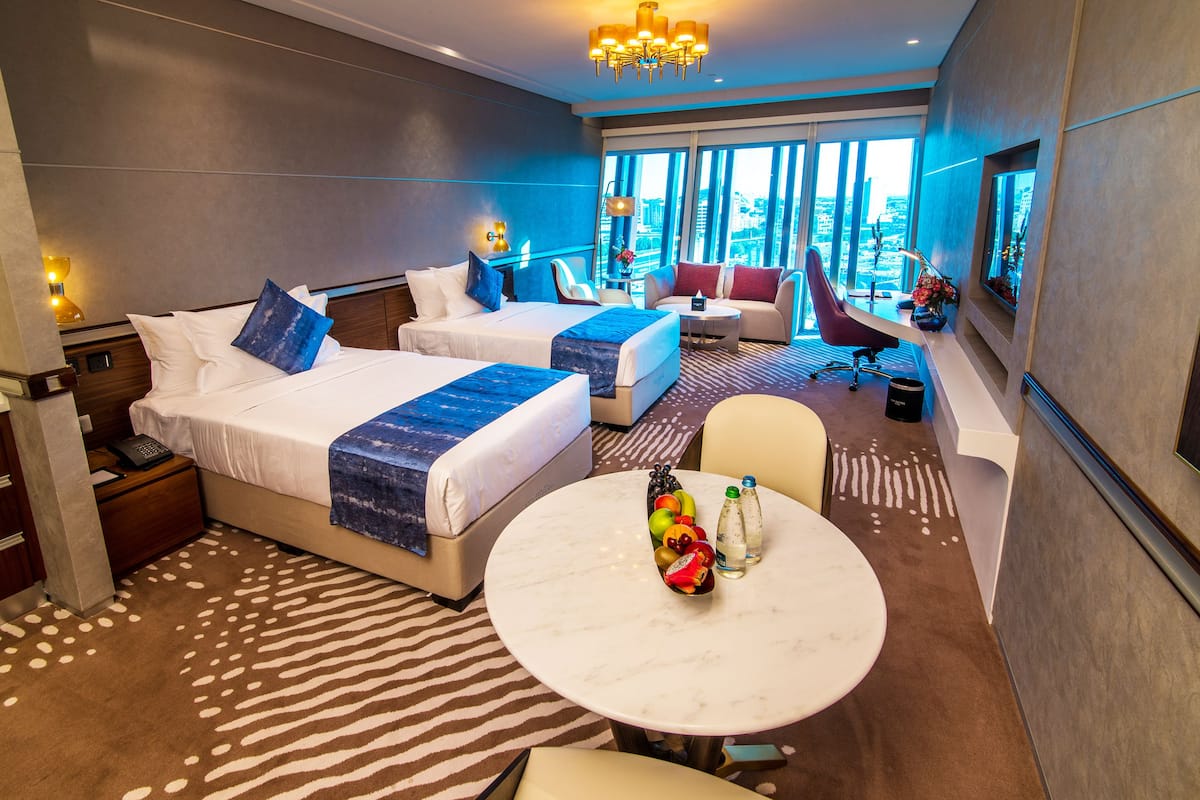 A hotel room with two beds, a sofa, a round table with fruit and bottles, and a city view.