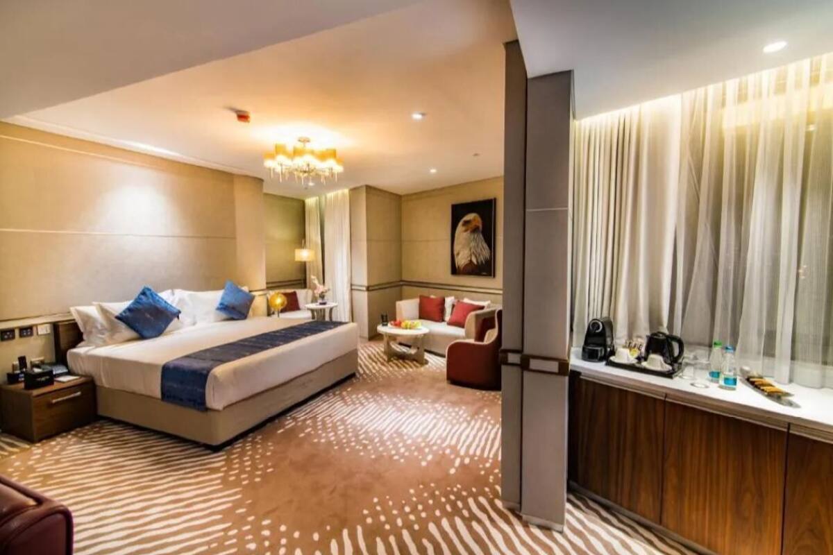 A modern hotel room with a large bed, a sitting area with a sofa and armchairs, a dining table, and a minibar.
