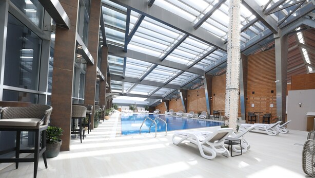 Una piscina interior con sillones de descanso y tumbonas, rodeada de amplios ventanales y un techo alto.