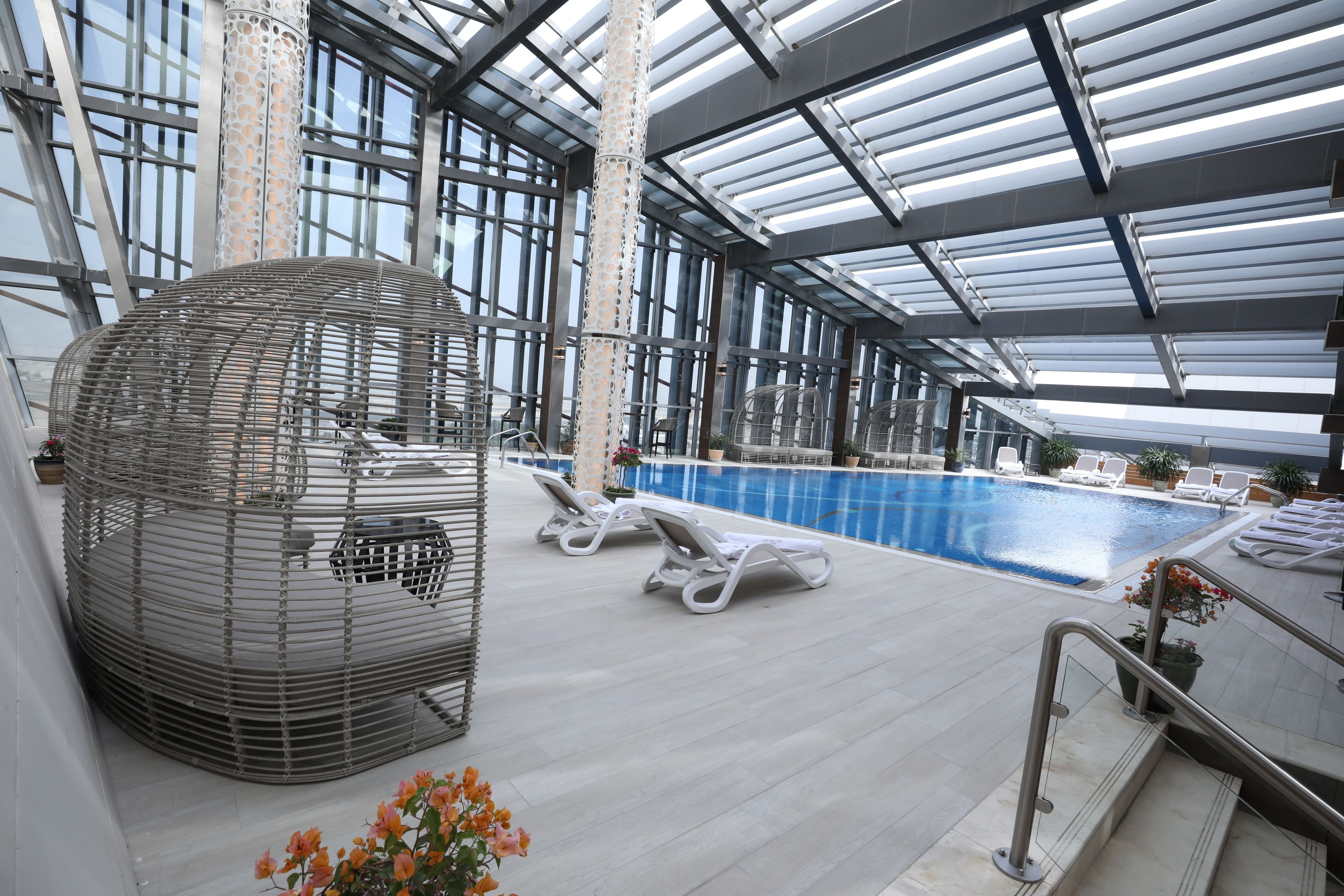 An indoor swimming pool with a glass ceiling, lounge chairs, and a decorative metal structure.