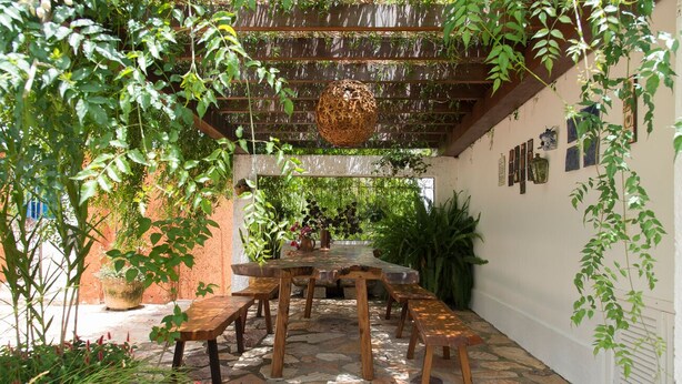 Un pati de pedra amb taula i bancs de fusta, envoltat de vegetació abundant i plantes en testos.
