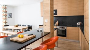 A modern kitchen with wooden cabinets, a black countertop, and a dining area with a wooden table and orange chairs. - Central Duplex Apartment with Large Terrace (Chamonix-Mont-Blanc)