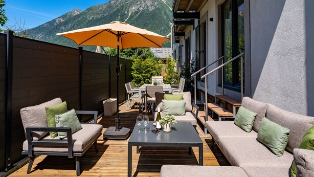 An outdoor seating area with a wooden deck, cushioned sofas, a table with wine glasses, and an umbrella. - Central Duplex Apartment with Large Terrace (Chamonix-Mont-Blanc)