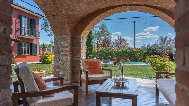 Eine Terrasse im Freien mit Holzmöbeln, einem Ziegelbogen und Blick auf ein Haus und einen Garten.