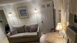 Un soggiorno con un divano beige, due cuscini, un tavolino rotondo in legno con una lampada e una porta bianca con un quadro sopra.