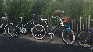 Fila di biciclette parcheggiate lungo un marciapiede, con un recinto bianco e vegetazione sullo sfondo.