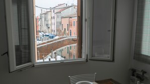 A dining area with a table set for two, a window with a view of a street and buildings, and a radiator on the wall. - Casa Bastianello large apartment, view of Canal Vena Historic Center comfort (Chioggia)