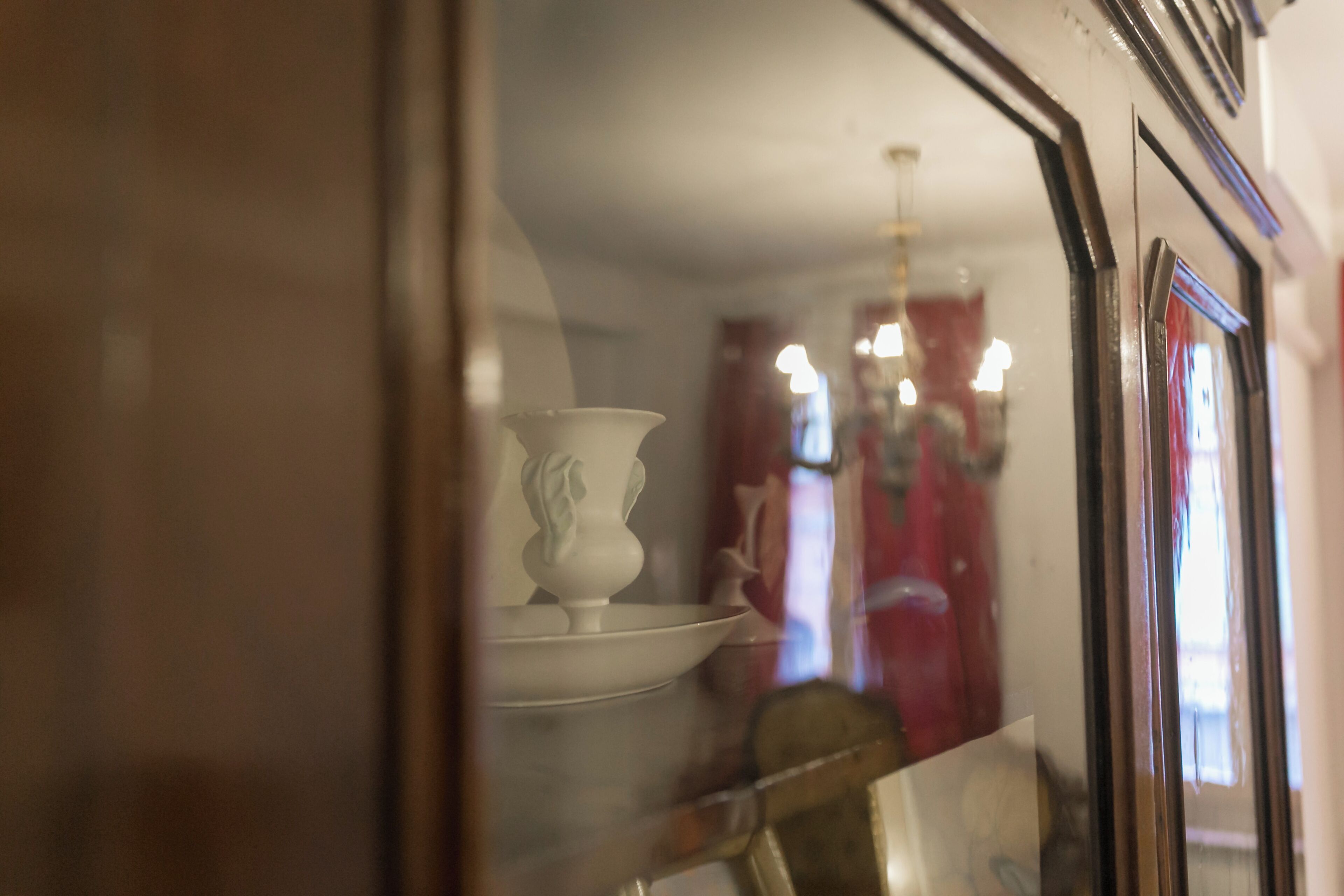 Une pièce avec un vase blanc posé sur une table, un miroir qui reflète une personne, et un rideau rouge en arrière-plan.
