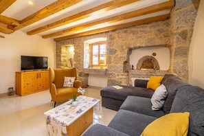 A living room with stone walls, a wooden ceiling, a TV, a sofa, a chair, and a small table with a vase. - Rustic holiday house HARMONIE private pool and large garden (Brusići)