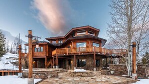 A large, multi-story wooden lodge with a stone foundation, multiple balconies, and a stone-paved entrance area. - Luxurious Hillside Lodge on a Working Wyoming Ranch (Little Jackson Hole)