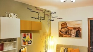Un salon comprenant un canapé, une lampe, un tableau encadré représentant le Colisée et un mur sur lequel sont inscrits des noms de villes.