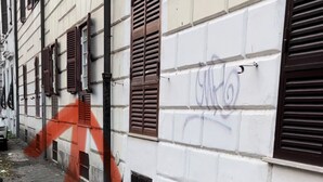 En smal brostensgade med en hvid bygning, der har brune skodder og et vindue med graffiti.