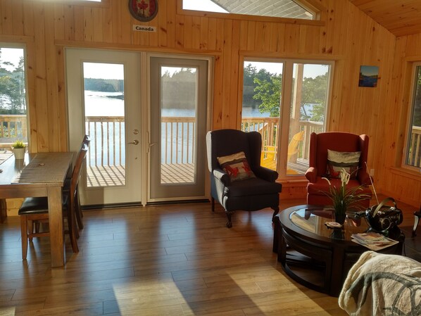 Interior de madera con una mesa de comedor, dos sillas, un sofá y vistas a un lago a través de puertas de vidrio.