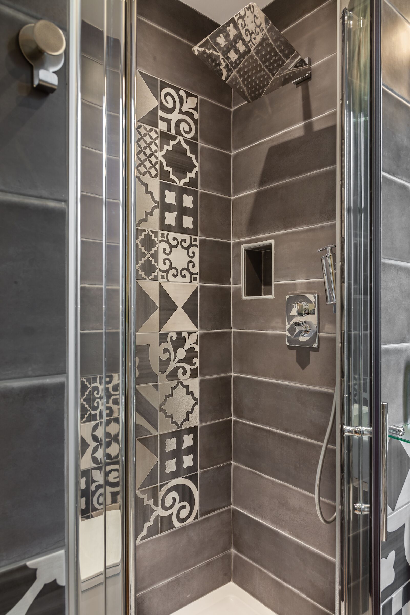 Una ducha con puerta de vidrio, azulejos decorados y una ventana pequeña cuadrada.