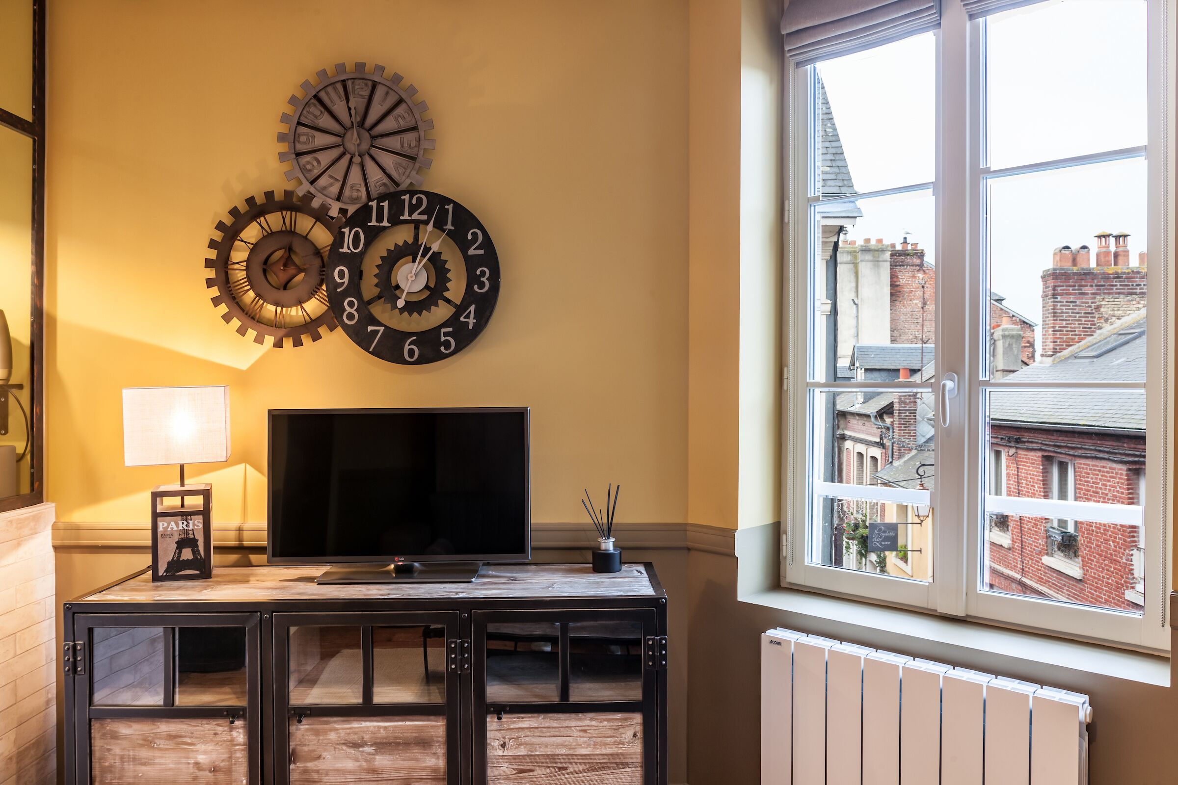 A room with a large window, a TV on a wooden cabinet, and two wall clocks.