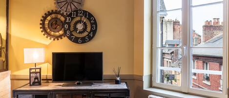 A room with a large window, a TV on a wooden cabinet, and two wall clocks.