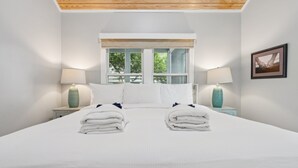 A neatly made bed with two towels, a window with a view of trees, and two lamps on nightstands. - Stones Throw | Walk to Beach & Center St | 2 Kings + Fenced Yard | Pet Friendly (Folly Beach)