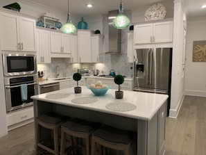 A modern kitchen with white cabinets, stainless steel appliances, a central island with stools, and decorative elements like a teal pendant light and a bowl of decorative balls.