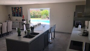 A modern kitchen with a large island, a dining area with chairs, a view of a swimming pool, and a kitchen counter with a toaster. - Villa overlooking the hill between Hyeres and St Tropez (Solliès-Pont)