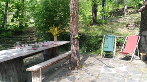 Una mesa de picnic con platos, vasos y un jarrón de flores en una zona boscosa con senderos de piedra y una silla roja.