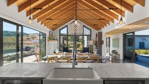 A modern kitchen with a large island, wooden ceiling beams, and large windows offering a view of the outdoors.