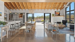 A spacious kitchen with a large island, a dining area with wooden chairs, and a view of the outdoors through large windows.